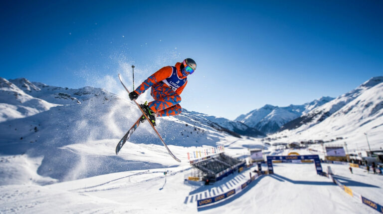 Le ski acrobatique aux Jeux olympiques d’hiver 2026 Un skieur acrobatique effectue un saut spectaculaire dans les airs sur fond de montagnes enneigées et un ciel bleu.